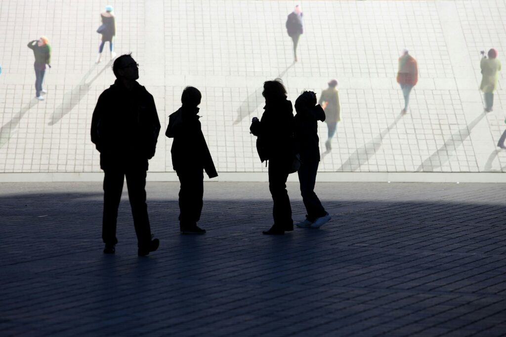 Silhouetted people walk on a sunny, paved area.